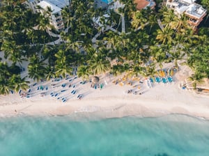 This is a beautiful beach scene captured from an aerial perspective. The sandy beach is dotted with numerous blue and yellow umbrellas providing shade for beachgoers.